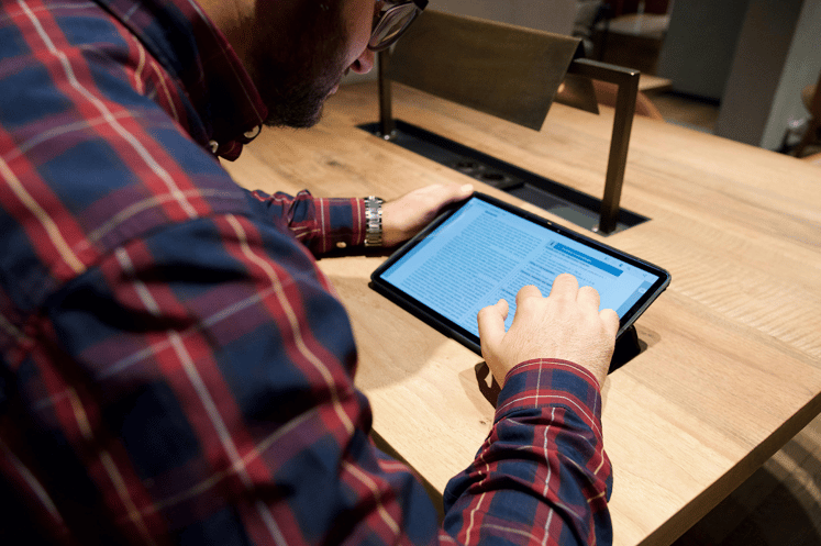 Man in a plaid shirt reading content on a tablet at a wooden desk.