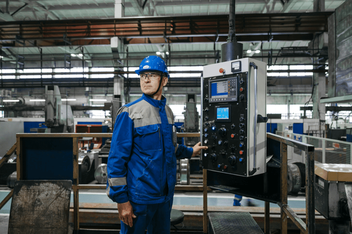 Factory worker in a blue uniform and a hard hat operating an industrial machine control panel.