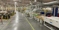 Interior of a large industrial manufacturing facility with machinery, conveyor lines, ducts, and storage drums.