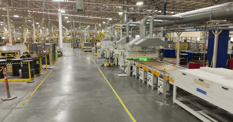 Interior of a large industrial manufacturing facility with machinery, conveyor lines, ducts, and storage drums.