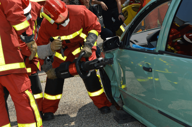Firefighters in orange gear use hydraulic rescue tools to cut open a damaged green car door.
