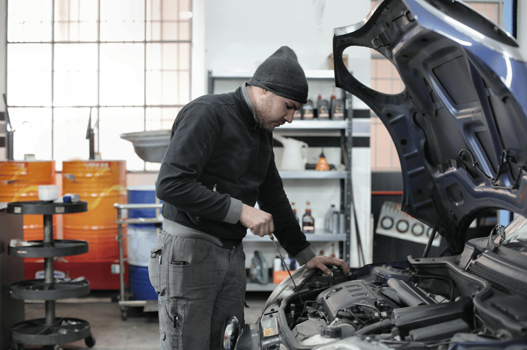Mechanic in a beanie checking under the hood of a blue car in an auto repair shop.