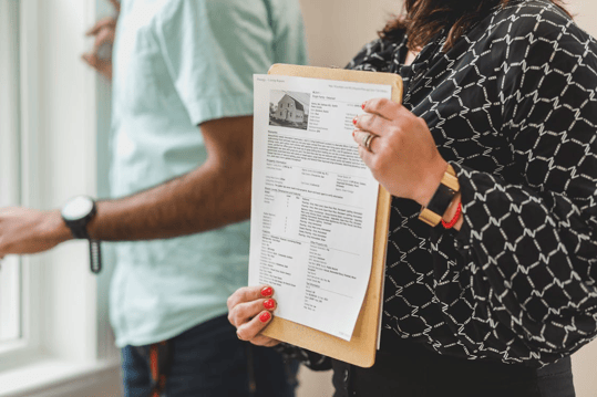 A person holding a document with a picture of a house and other information on it, with another person in a light blue shirt in the background. 