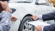 Two people inspect damage on a white car, pointing at scratches above the front wheel, while one holds a pen and an insurance form on a clipboard.