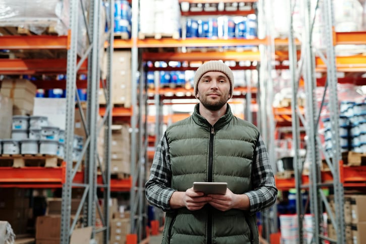 Man in a beanie and a vest holding a tablet in a warehouse with orange shelving.