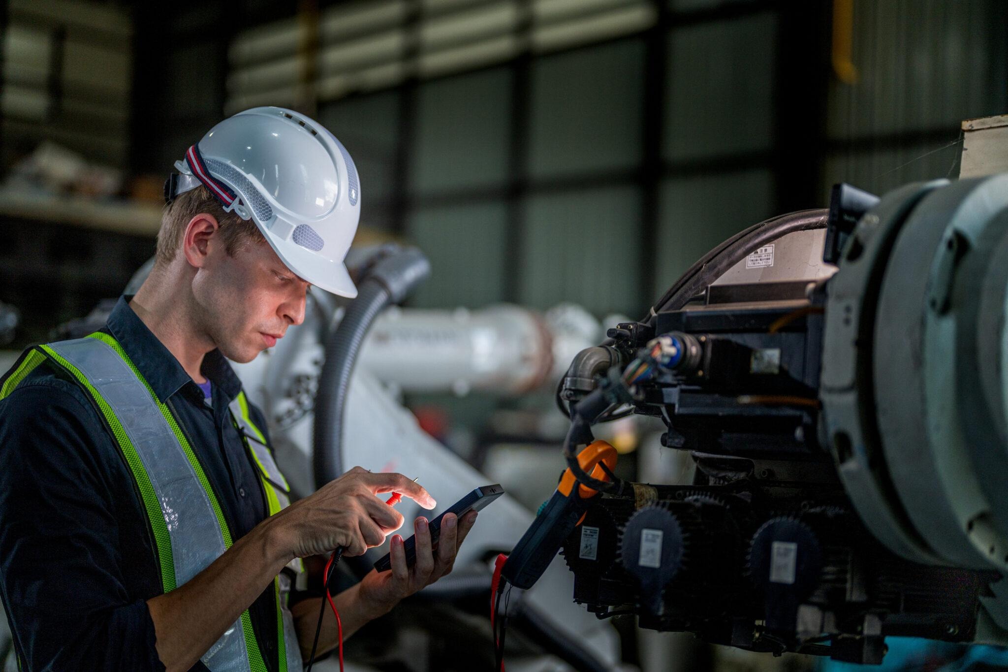 An engineer in a hard hat and safety vest is inspecting industrial machinery with a diagnostic tool.