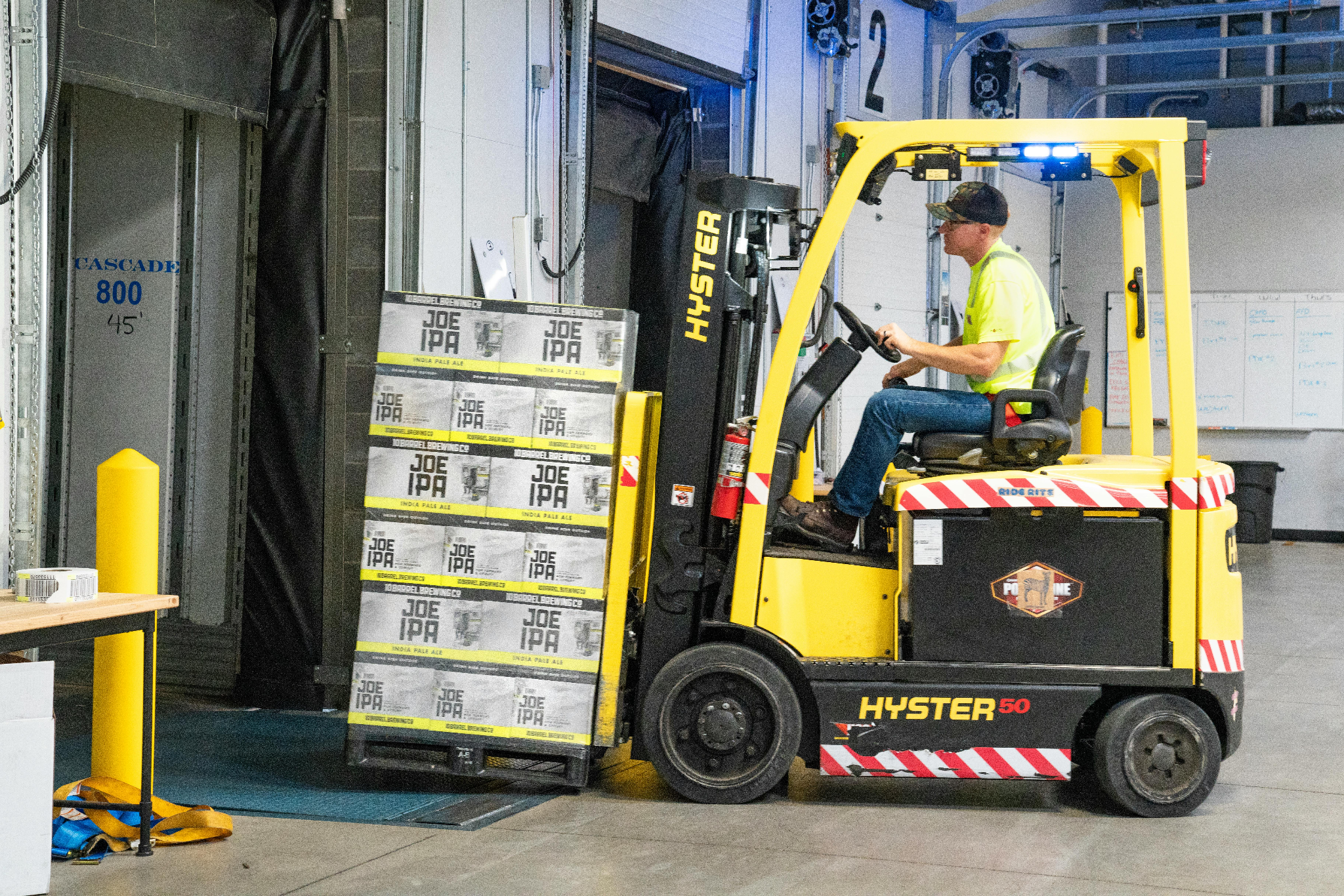 Worker in a high-visibility vest operating a yellow Hyster forklift carrying stacked cases in a warehouse