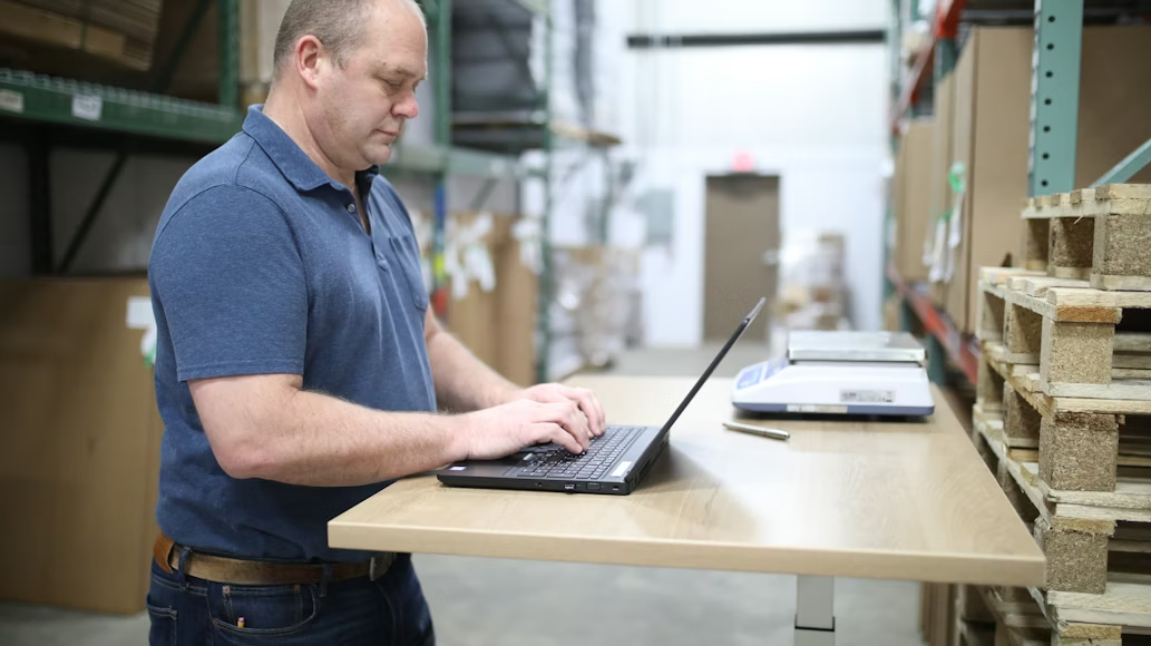 Man in a dark blue top and jean trousers working on a laptop standing between warehouse storage shelves