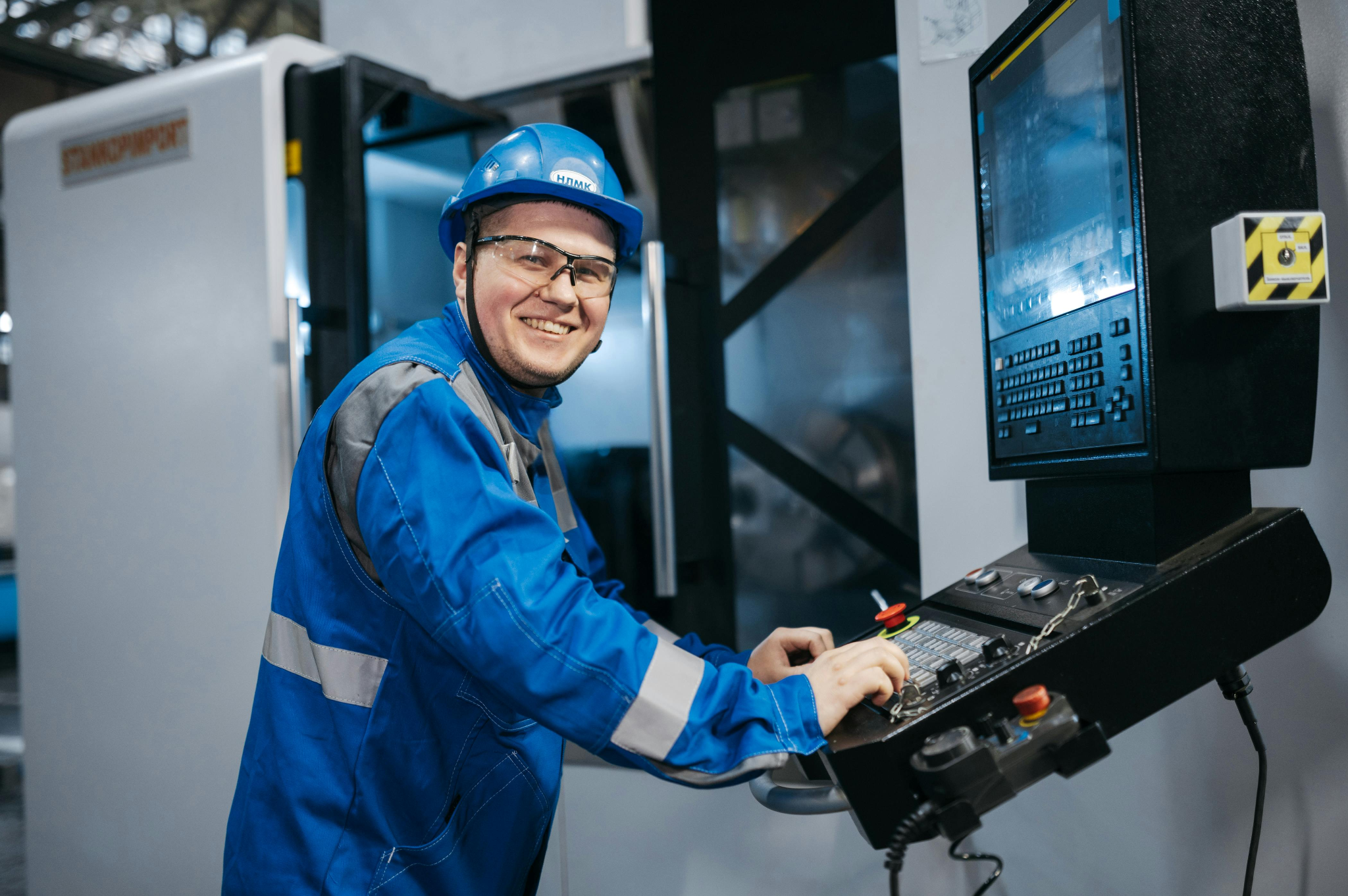Smiling factory worker in blue uniform and hard hat operating a CNC machine control panel.