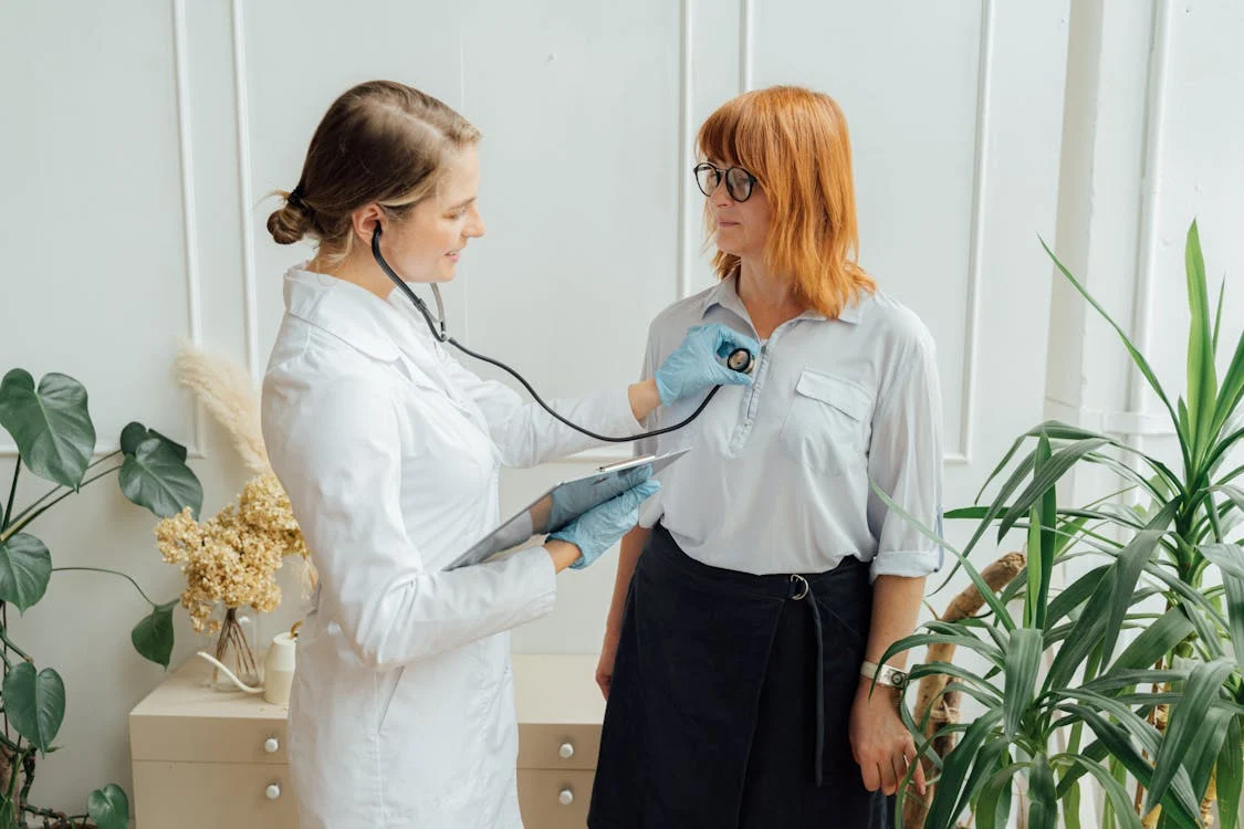 Healthcare professional using a stethoscope to examine a patient in a medical office and documenting it digitally on a tablet