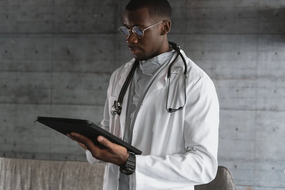 A doctor in a white coat, holding a tablet, stands in a professional, modern clinical setting