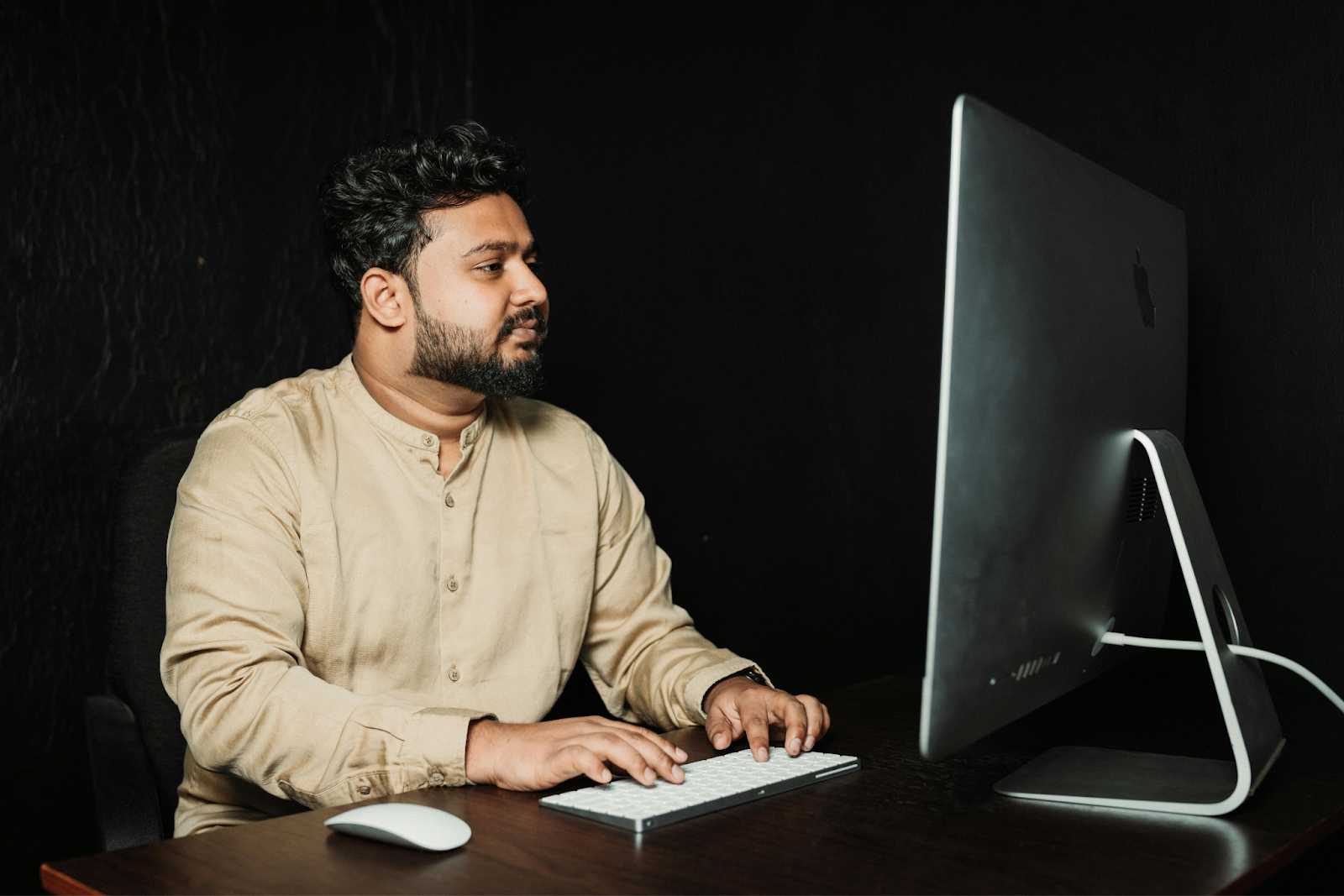 Man in beige shirt working at desktop computer with keyboard and mouse against a dark background.