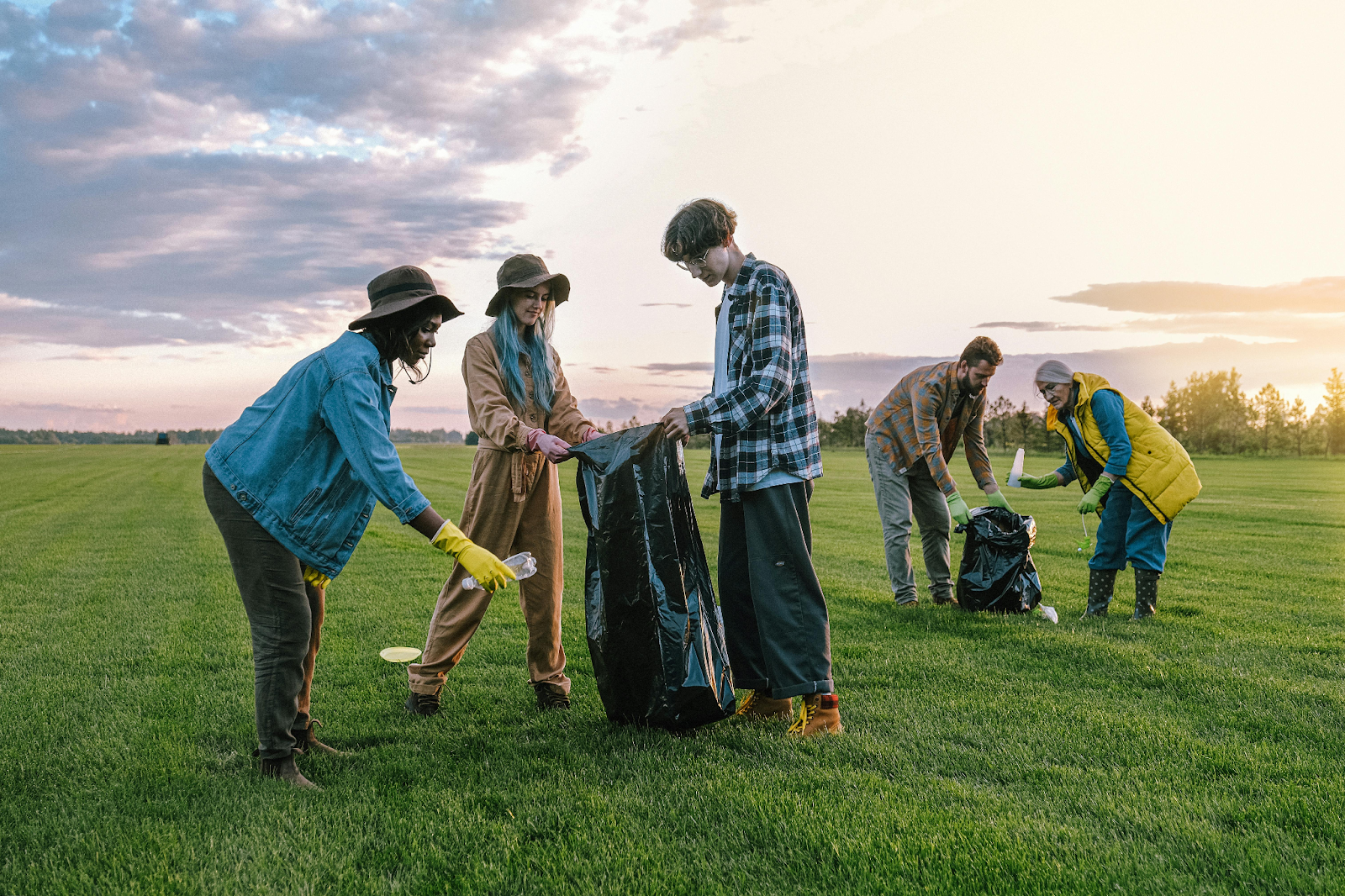 A Group of five volunteers picking up litter with garbage bags in a grassy field at sunset.