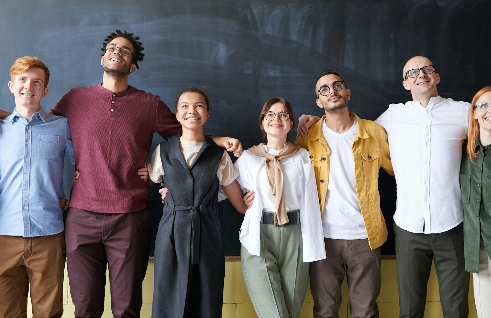 Group of seven smiling people standing together with arms around each other in front of a chalkboard.