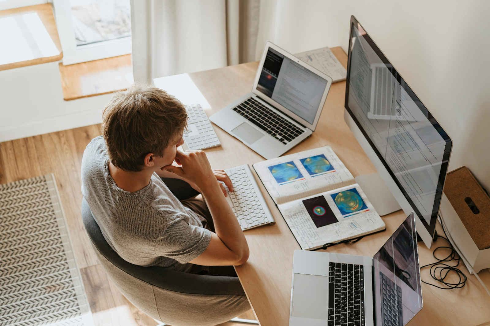 Person working at home office desk with dual monitors, laptops, and notes.