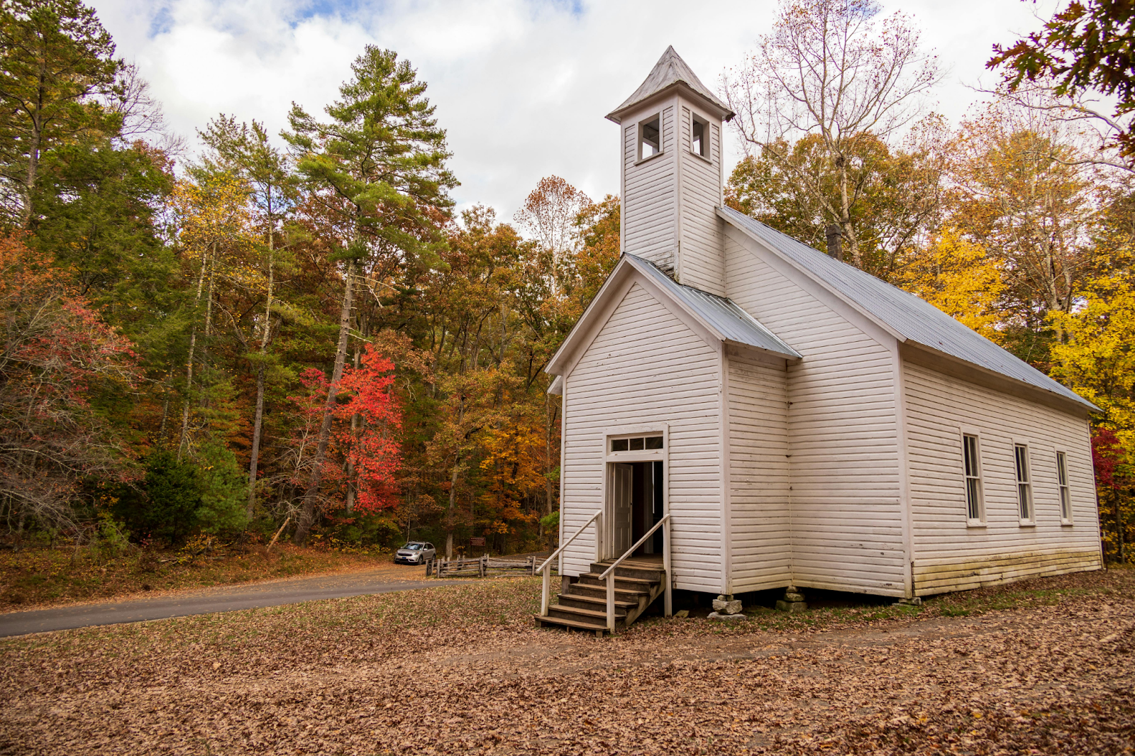Small white wooden church with steeple surrounded by colorful autumn foliage in a rural setting.