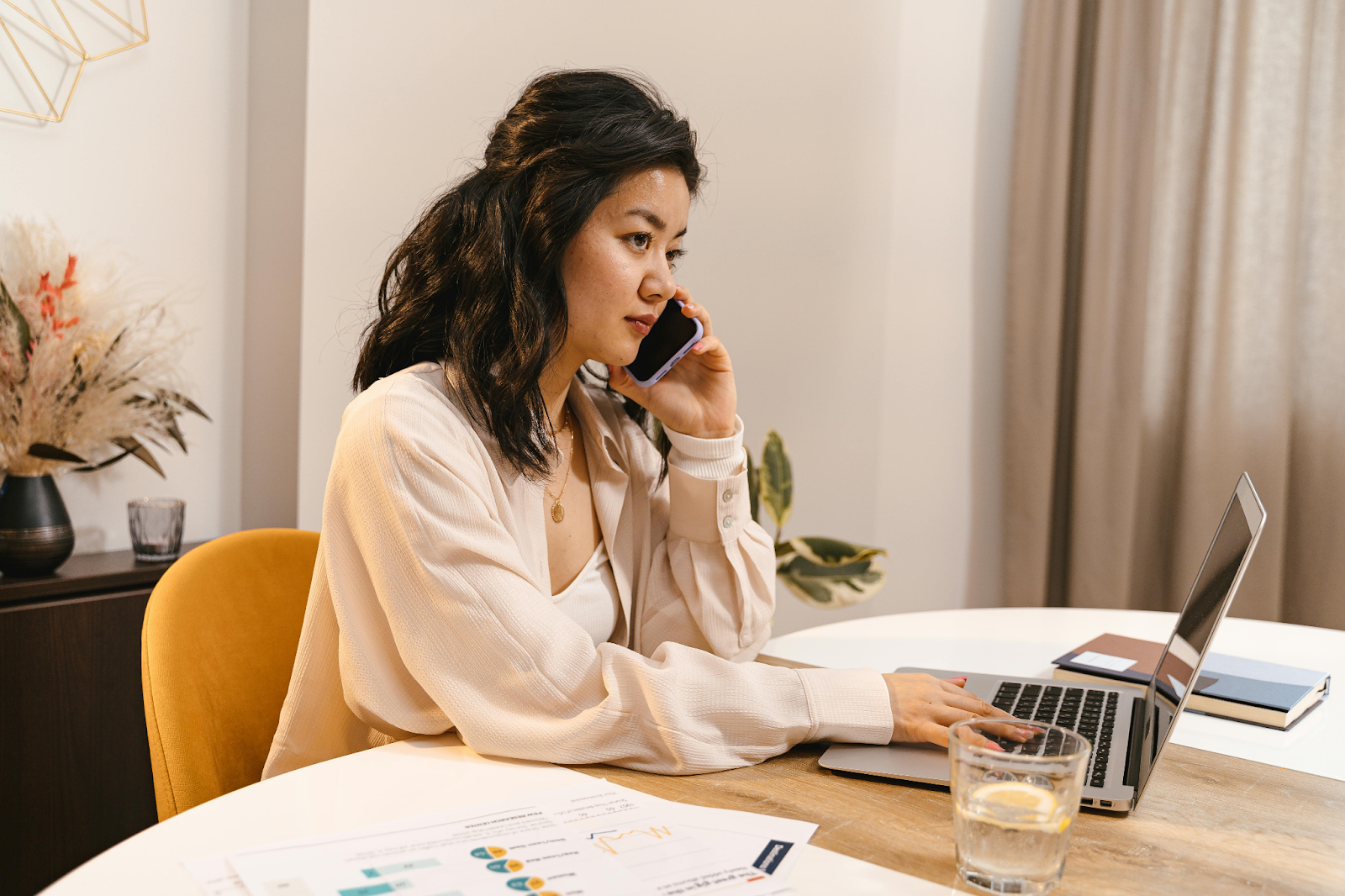 Woman talks on phone while working on laptop at desk with documents and a glass of water.
