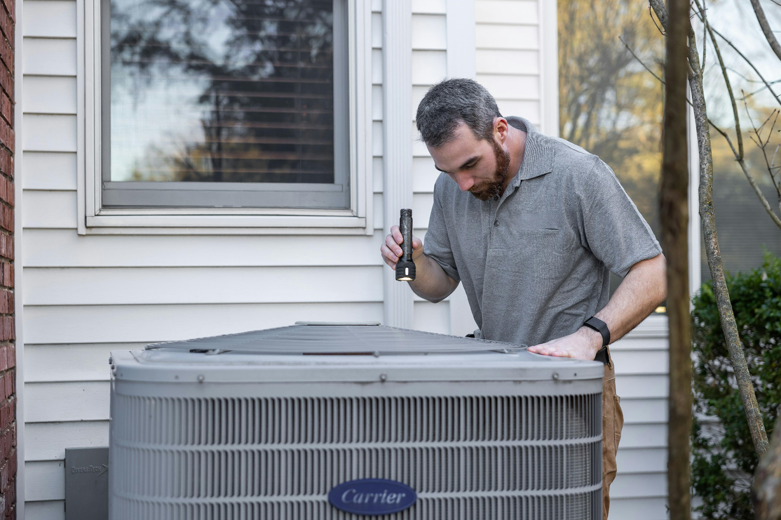 Technician using a flashlight to inspect an outdoor Carrier air conditioning condenser unit beside a house