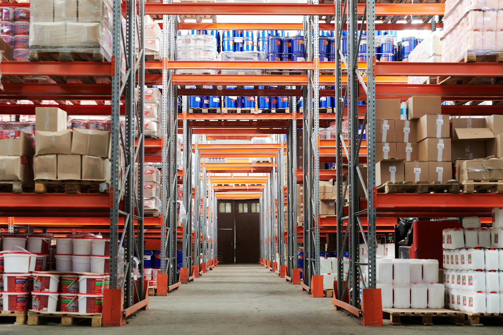 Long warehouse aisle with orange steel shelving stocked with boxes, blue drums, and plastic containers.