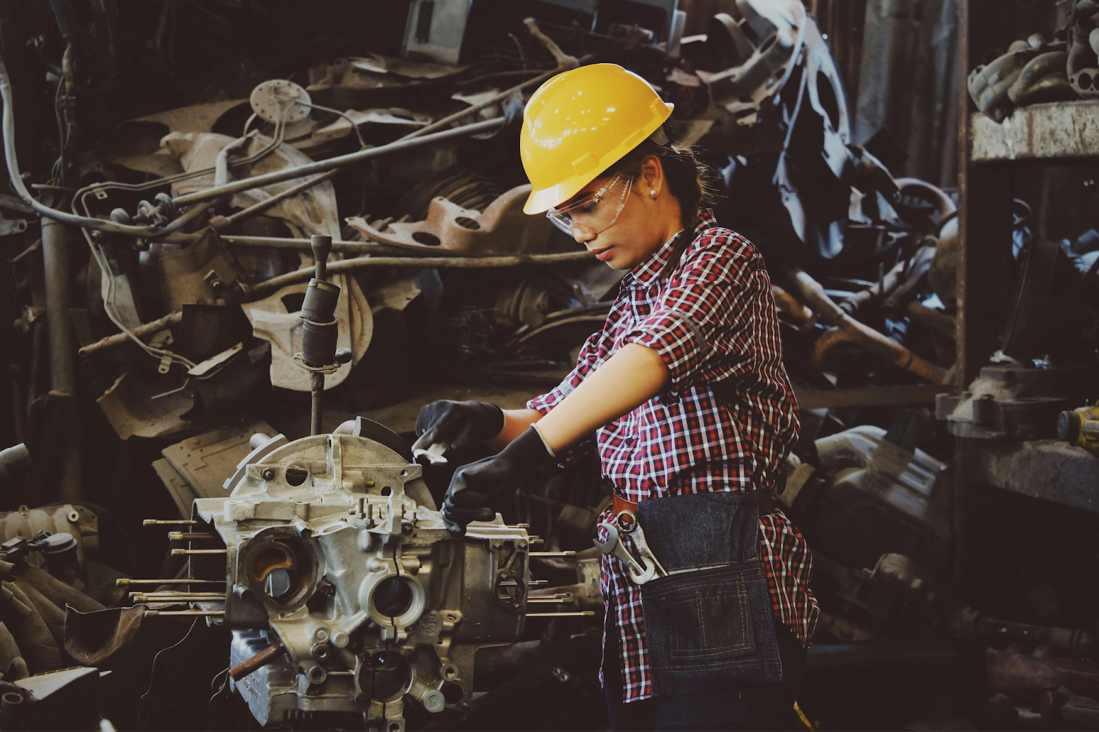 Woman in a yellow hard hat and safety glasses working on an engine in a mechanical workshop.