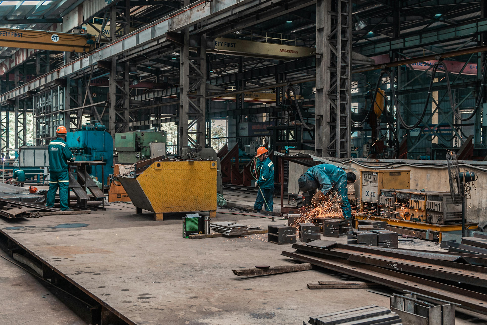 Workers in blue uniforms cutting and grinding metal inside a large industrial steel fabrication workshop.