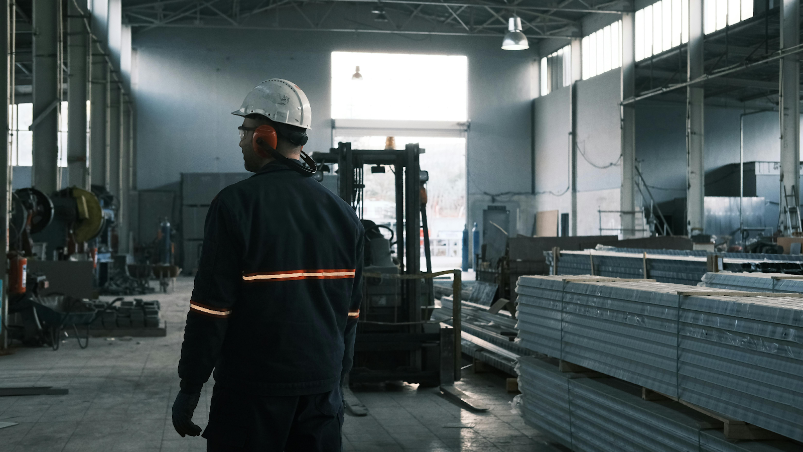 Worker in a hard hat and high-visibility jacket walking through an industrial warehouse with a forklift nearby.