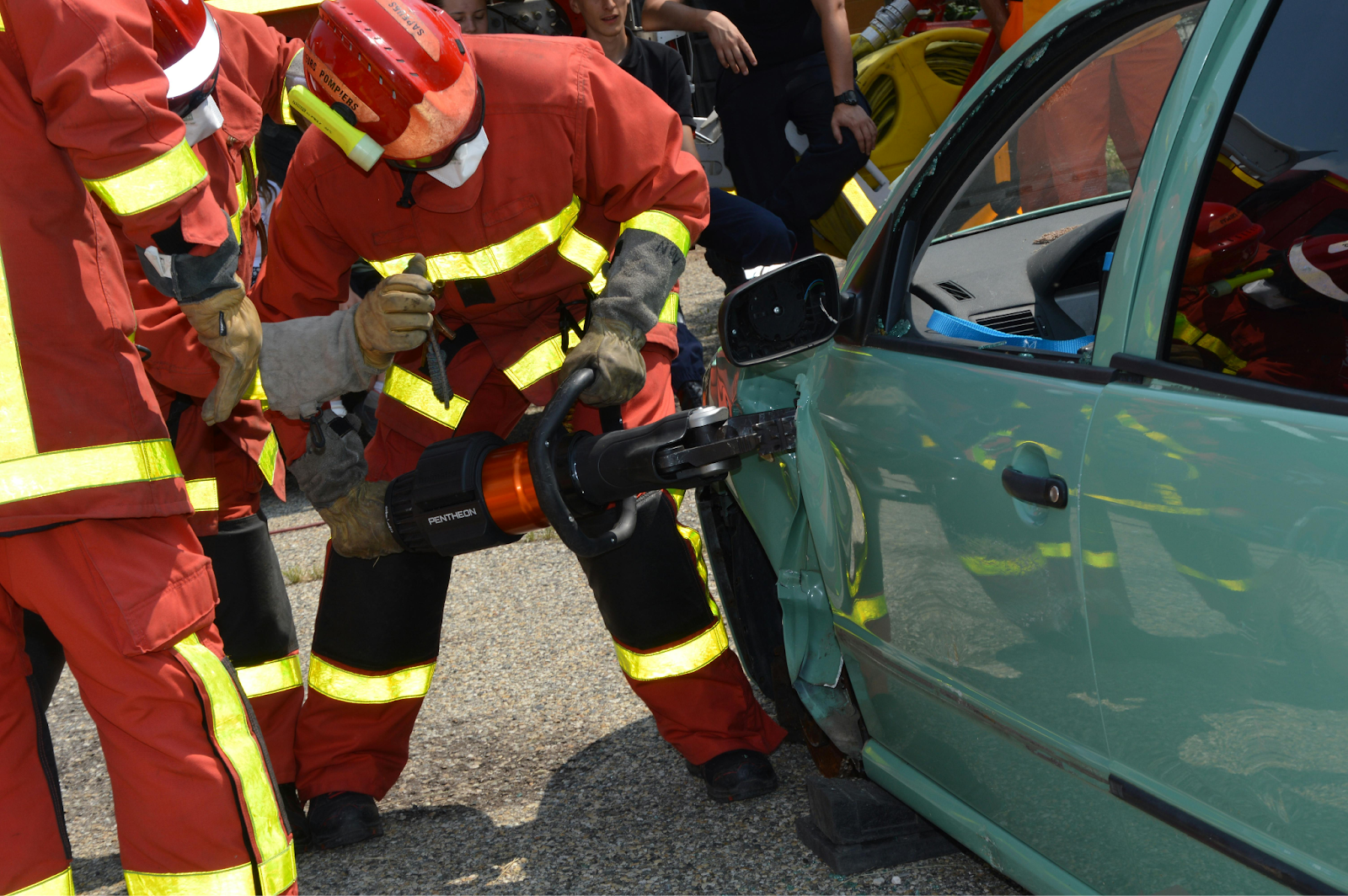 Firefighters in orange gear use hydraulic rescue tools to cut open a damaged green car door.