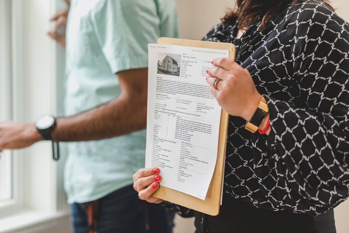 A person holding a document with a picture of a house and other information on it, with another person in a light blue shirt in the background
