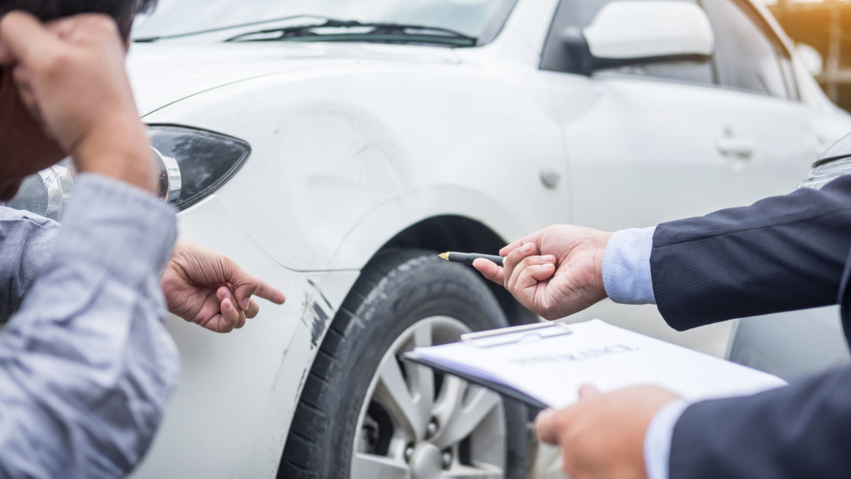 Two people inspect damage on a white car, pointing at scratches above the front wheel, while one holds a pen and an insurance form on a clipboard.