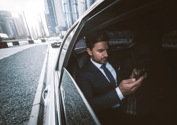 Businessman in a suit sitting in the backseat of a limousine, looking at his smartphone while traveling through a modern city with tall buildings.